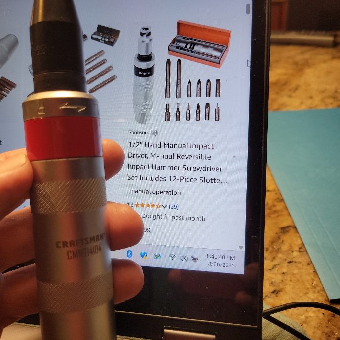 A hand holding an impact screwdriver. Behind it is a laptop showing an Amazon product: 1/2